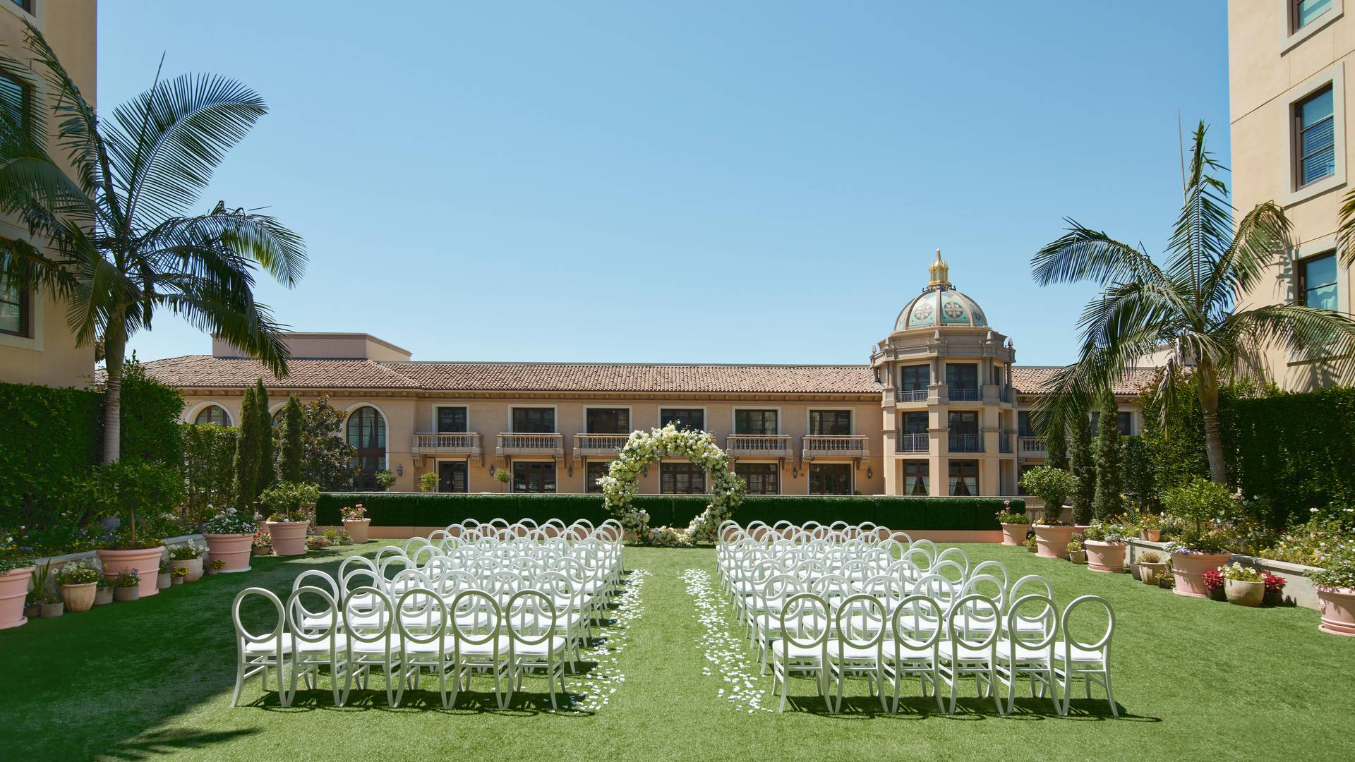 A picturesque outdoor ceremony setup with rows of white chairs arranged on lush green lawns, framed by Mediterranean-style architecture and swaying palm trees under a clear blue sky.
