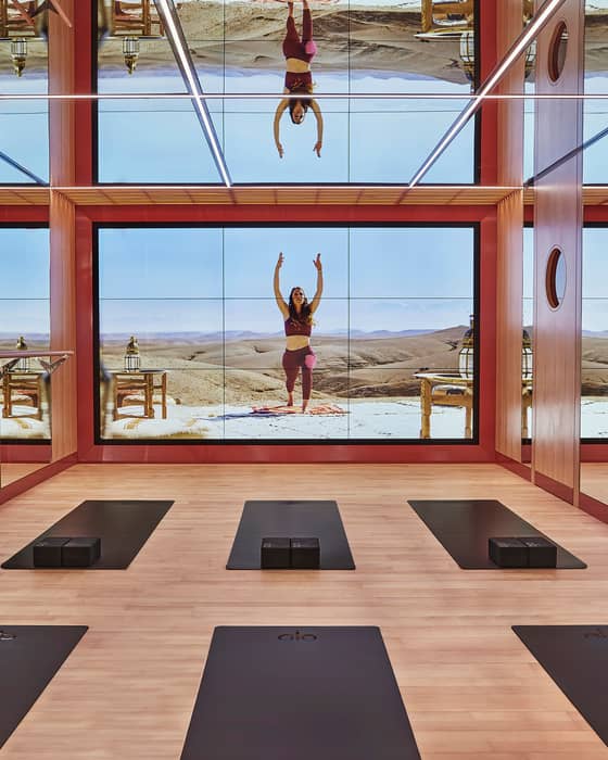 A tranquil yoga room with mirrored walls, wooden flooring, and a digital projection of a desert wellness retreat.