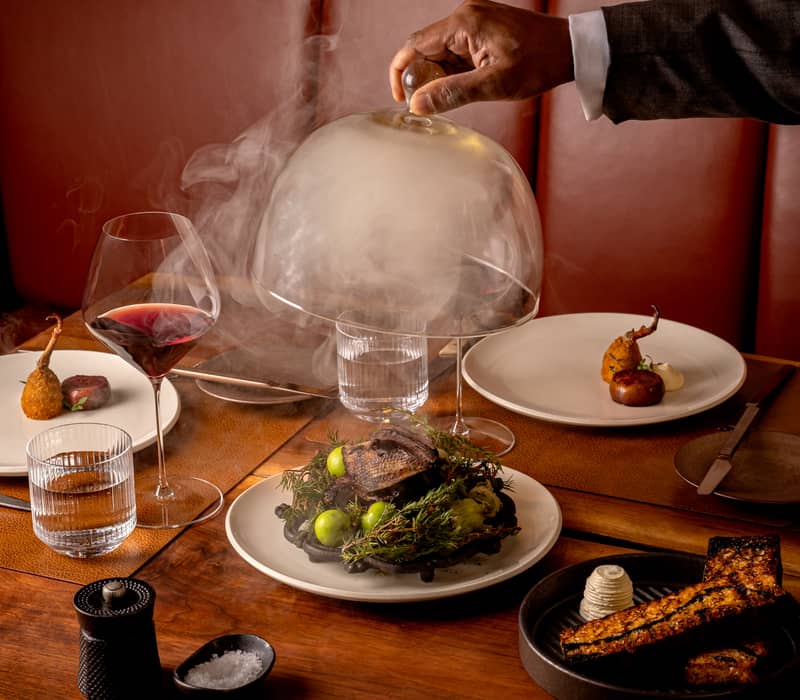 Luxurious dining setup featuring a smoked entrée unveiled under a glass cloche, paired with fine wine and artistic sides.