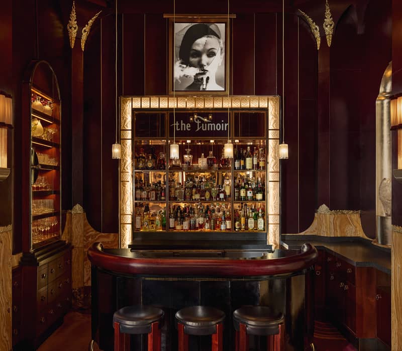 Decadent cocktail bar with Art Deco details, rich burgundy tones, illuminated shelving, and an intimate ambiance framed by soft lighting