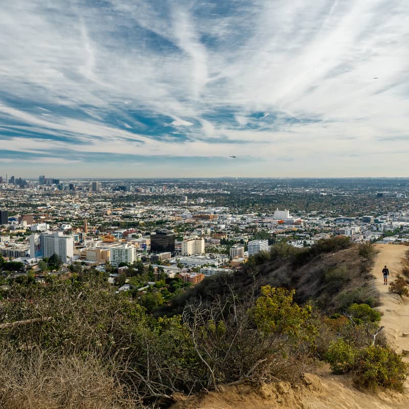 Hikers on a dirt trail overlooking Los Angeles skyline under dramatic, streaked clouds.