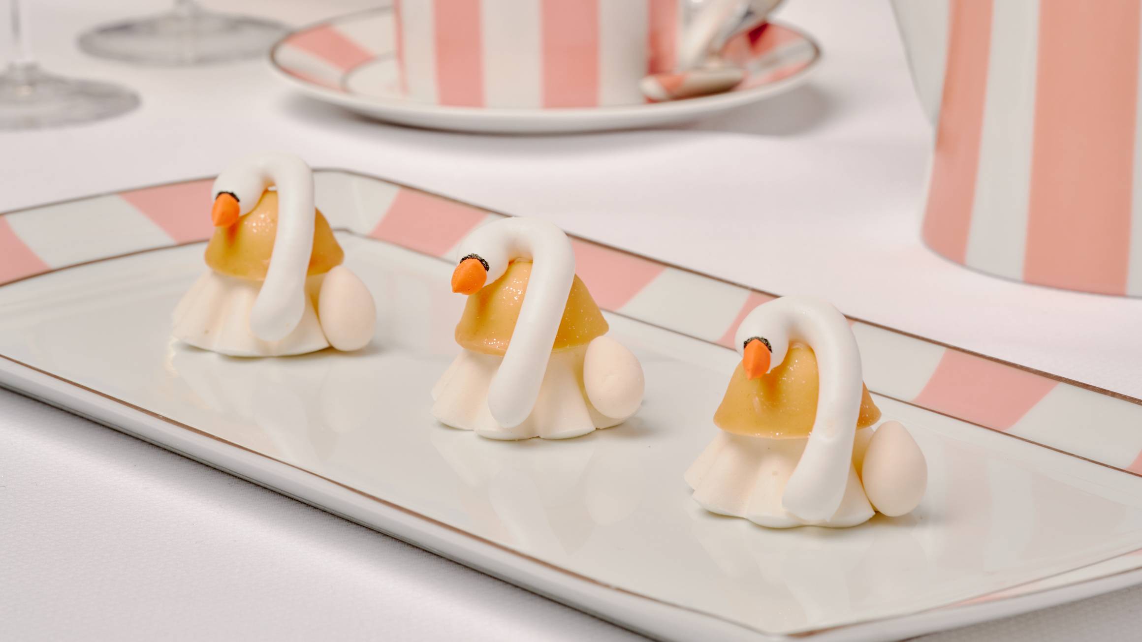 Three delicate swan-shaped petit fours made with white and golden elements, placed on a striped plate with matching pink-and-white teacup and teapot in the background.
