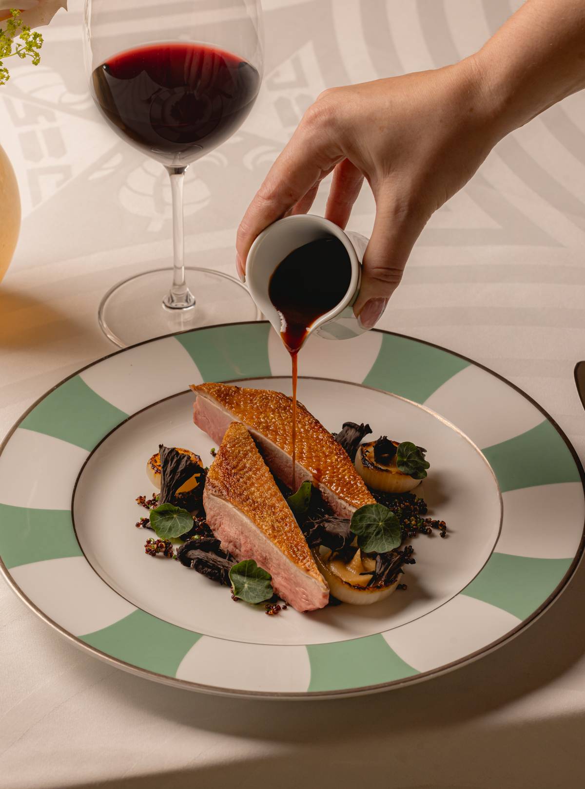 A plated duck breast dish, garnished with greens and mushrooms, accompanied by a glass of red wine and rich sauce