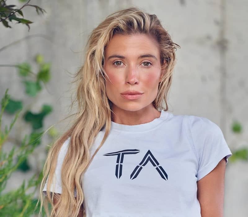 Tracy Anderson, with long blonde hair, wearing a white T-shirt featuring a bold "TA" logo, against a natural, leafy background