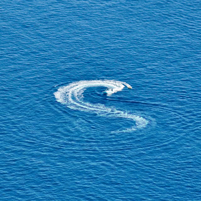 A speedboat creates a striking "S" pattern of white wake across the vibrant, endless expanse of deep blue ocean.