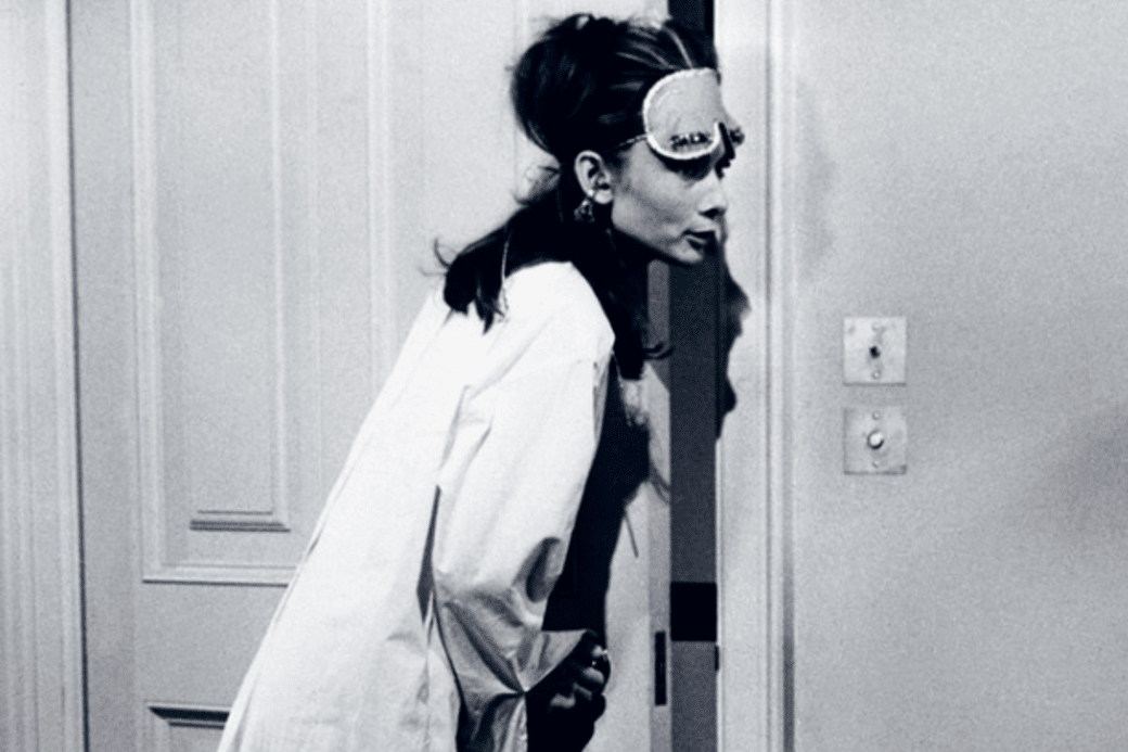 A woman wearing a sleep mask and oversized shirt leans curiously through a partially opened door in a dimly lit room.